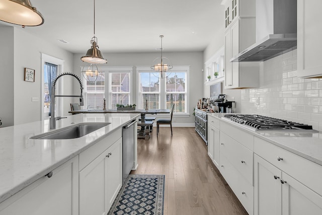 kitchen featuring wood finished floors, stainless steel appliances, a sink, white cabinets, and wall chimney range hood