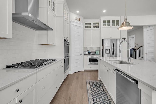 kitchen with wall chimney range hood, decorative light fixtures, appliances with stainless steel finishes, light wood-style floors, and a sink