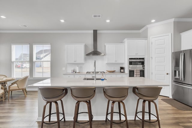 kitchen featuring wall chimney exhaust hood, white cabinetry, an island with sink, stainless steel appliances, and light hardwood / wood-style floors