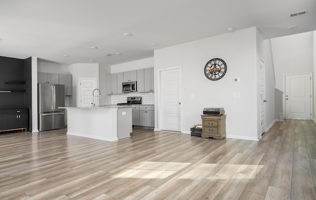 kitchen with decorative backsplash, light stone counters, gray cabinetry, stainless steel appliances, and an island with sink