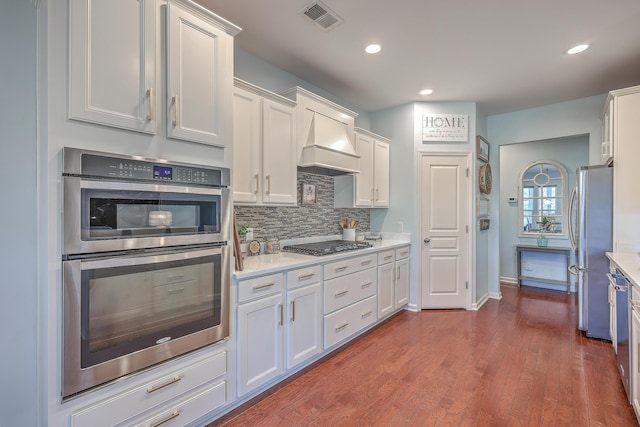 kitchen featuring appliances with stainless steel finishes, white cabinets, dark hardwood / wood-style flooring, decorative backsplash, and custom exhaust hood