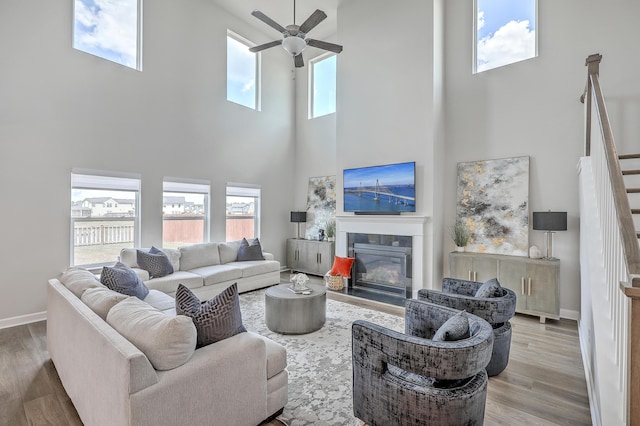 living room featuring a ceiling fan, a towering ceiling, wood finished floors, stairway, and a glass covered fireplace