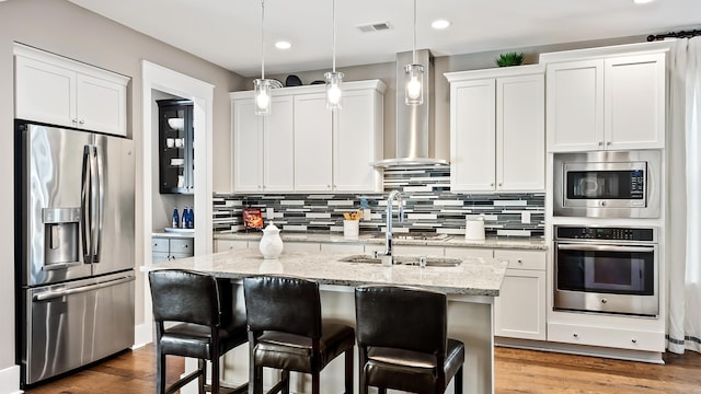 kitchen featuring appliances with stainless steel finishes, decorative light fixtures, white cabinetry, a kitchen island with sink, and wall chimney range hood