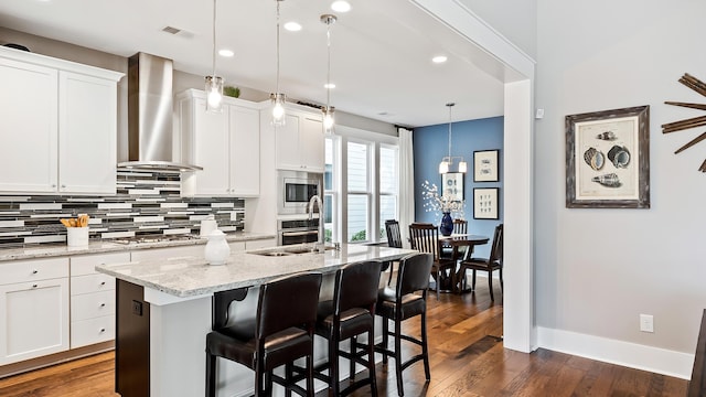 kitchen featuring an island with sink, appliances with stainless steel finishes, pendant lighting, and wall chimney exhaust hood