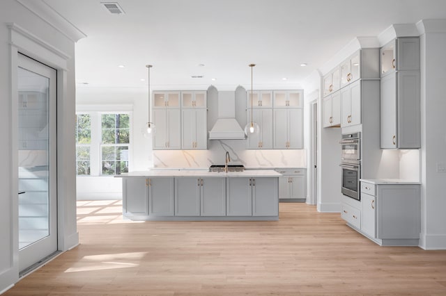 kitchen featuring gray cabinetry, custom exhaust hood, pendant lighting, a center island with sink, and light hardwood / wood-style flooring