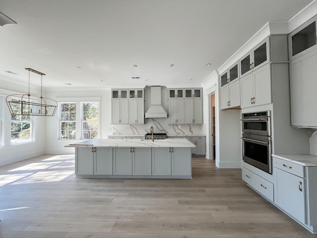 kitchen with light stone countertops, premium range hood, stainless steel double oven, pendant lighting, and a center island with sink