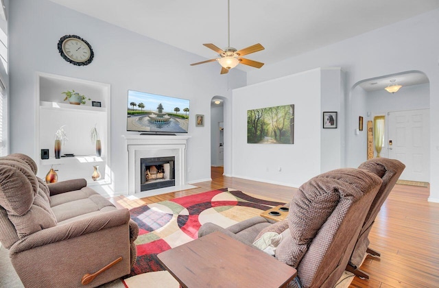 living area featuring ceiling fan, baseboards, a fireplace with flush hearth, light wood-type flooring, and arched walkways