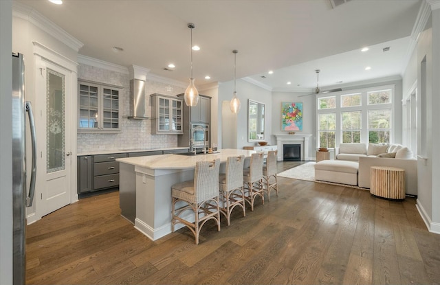 kitchen with wall chimney exhaust hood, a center island with sink, gray cabinets, stainless steel appliances, and decorative backsplash