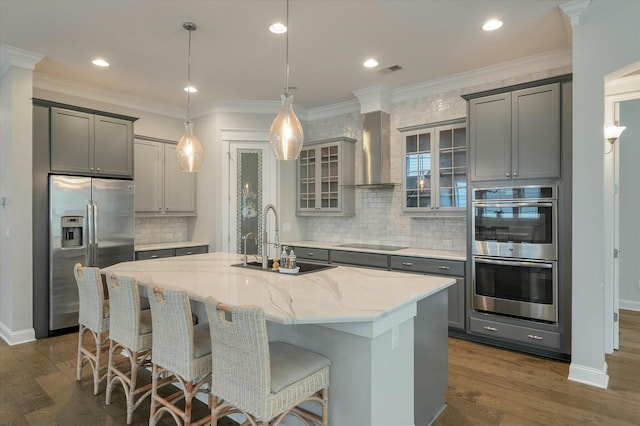 kitchen featuring wall chimney exhaust hood, stainless steel appliances, a center island with sink, and gray cabinetry