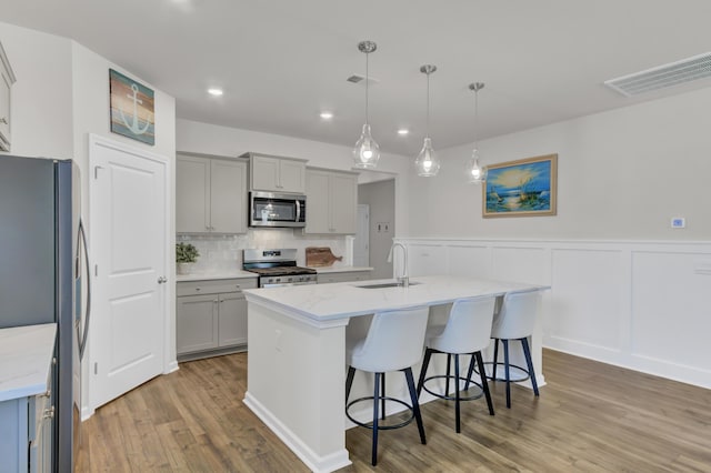 kitchen featuring a kitchen island with sink, appliances with stainless steel finishes, hanging light fixtures, gray cabinetry, and sink