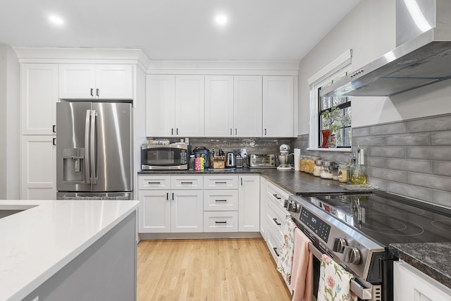 kitchen featuring appliances with stainless steel finishes, white cabinetry, wall chimney range hood, light wood-type flooring, and backsplash