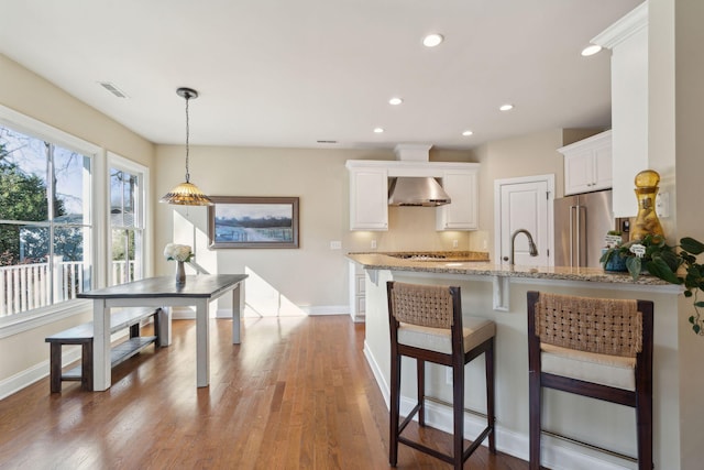 kitchen featuring light stone counters, a peninsula, white cabinets, high end fridge, and wall chimney range hood