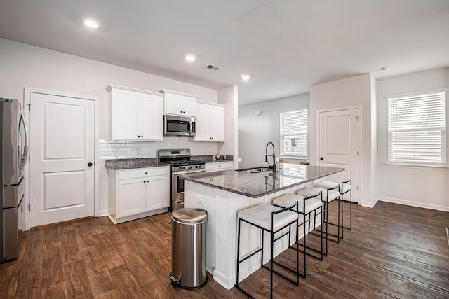 kitchen with stainless steel appliances, a kitchen island with sink, dark stone countertops, and white cabinets