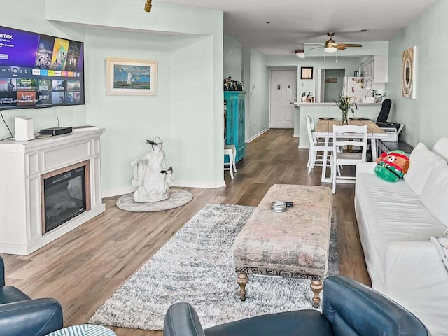 living area with a glass covered fireplace, ceiling fan, baseboards, and wood finished floors