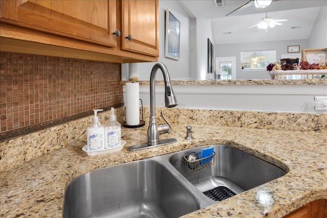 room details featuring ceiling fan, light stone counters, decorative backsplash, brown cabinetry, and a sink