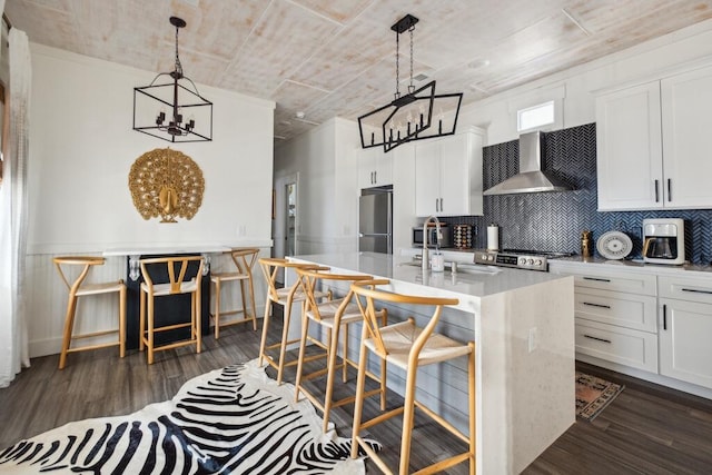 kitchen featuring wall chimney range hood, a chandelier, appliances with stainless steel finishes, a kitchen island with sink, and light countertops