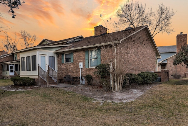 back of house at dusk featuring entry steps, a sunroom, a chimney, a yard, and brick siding