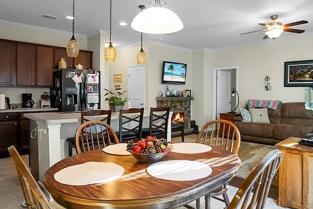 dining space with a stone fireplace, ceiling fan, crown molding, and light tile patterned flooring