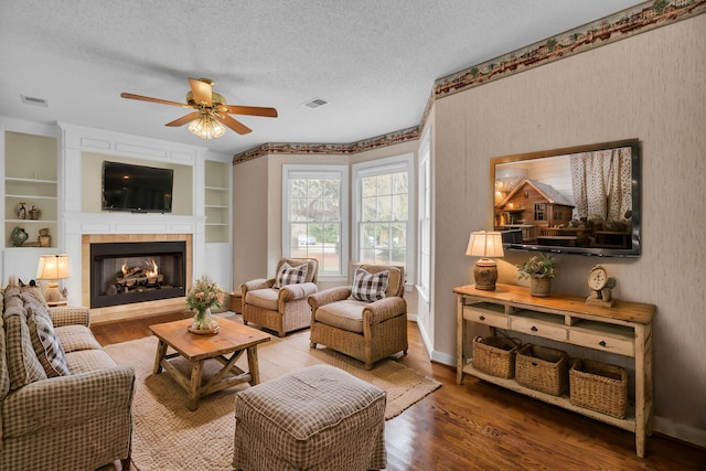 living room featuring a tile fireplace, ceiling fan, built in features, a textured ceiling, and wood-type flooring