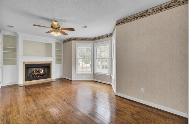 unfurnished living room with ceiling fan, built in features, wood-type flooring, a textured ceiling, and a tiled fireplace