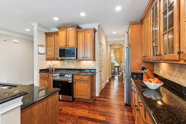 kitchen with stainless steel appliances, arched walkways, brown cabinetry, dark stone countertops, and ornamental molding