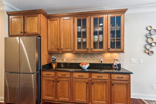 kitchen with freestanding refrigerator, decorative backsplash, dark stone countertops, glass insert cabinets, and brown cabinets