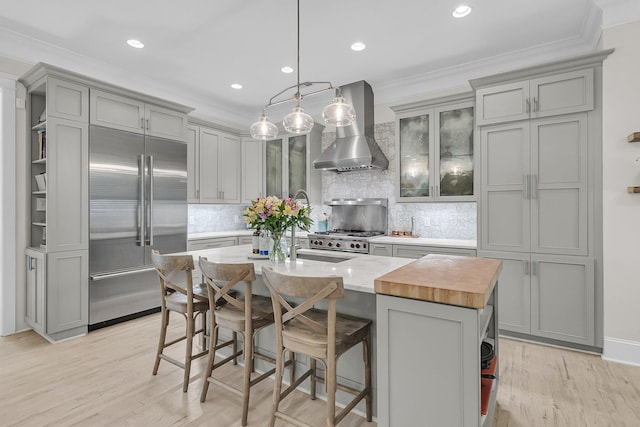 kitchen with wall chimney range hood, built in fridge, gray cabinets, ornamental molding, and light wood finished floors