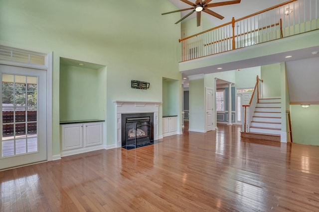 unfurnished living room featuring wood finished floors, a fireplace with flush hearth, ceiling fan, stairs, and a high ceiling