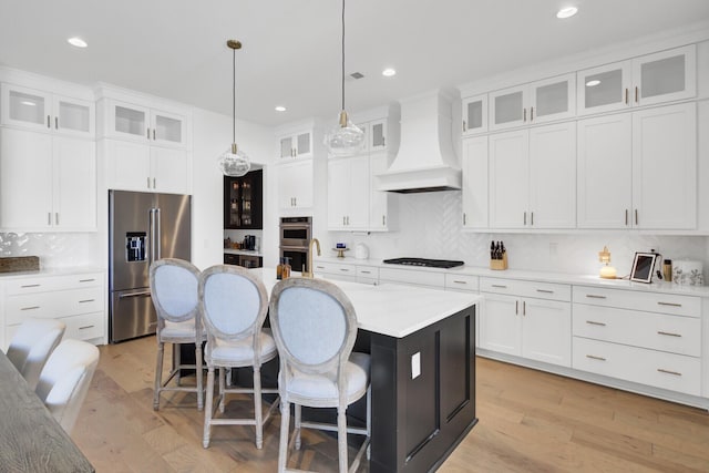 kitchen with light wood finished floors, a center island with sink, custom range hood, a kitchen breakfast bar, and stainless steel appliances