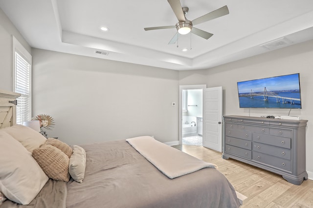 bedroom with light wood-type flooring, ceiling fan, a raised ceiling, and ensuite bath