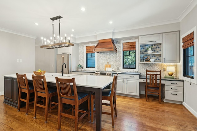 kitchen with stainless steel appliances, custom range hood, backsplash, light wood-style floors, and a kitchen island with sink