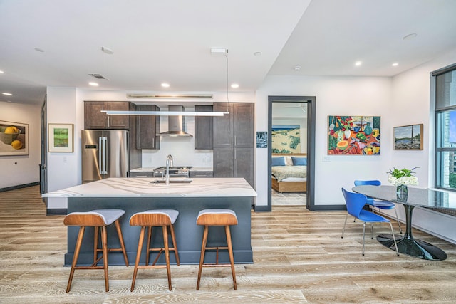 kitchen featuring a breakfast bar, high end refrigerator, a kitchen island with sink, dark brown cabinets, and wall chimney exhaust hood