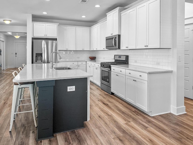 kitchen with stainless steel appliances, white cabinetry, an island with sink, light stone countertops, and light wood-type flooring