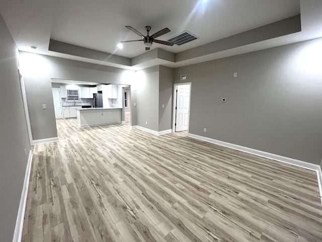 unfurnished living room featuring a raised ceiling, ceiling fan, and light hardwood / wood-style floors