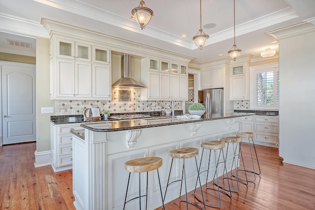 kitchen with a center island with sink, a raised ceiling, wall chimney exhaust hood, a kitchen breakfast bar, and stainless steel appliances