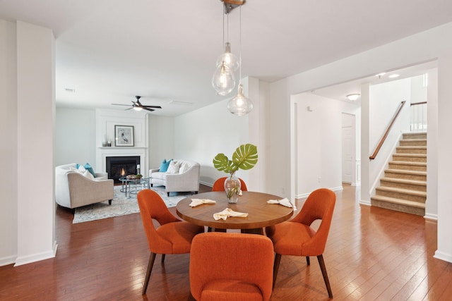 dining area featuring dark wood finished floors, a ceiling fan, a large fireplace, and stairway
