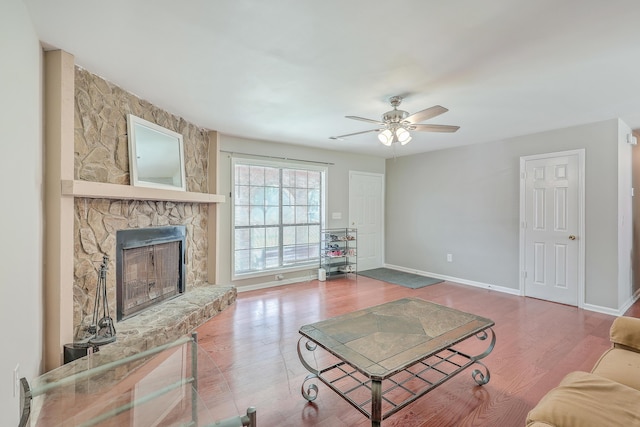 living room featuring a stone fireplace, wood finished floors, baseboards, and ceiling fan