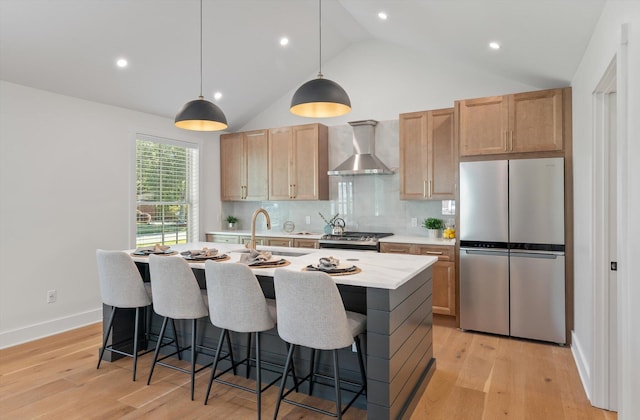 kitchen with wall chimney range hood, hanging light fixtures, an island with sink, light hardwood / wood-style floors, and stainless steel appliances