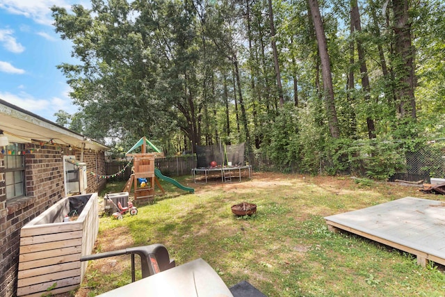 view of yard featuring a wooden deck, a trampoline, a fenced backyard, a playground, and a fire pit