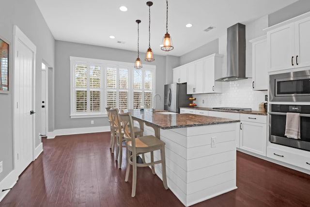 kitchen featuring wall chimney range hood, a kitchen island with sink, white cabinetry, and appliances with stainless steel finishes