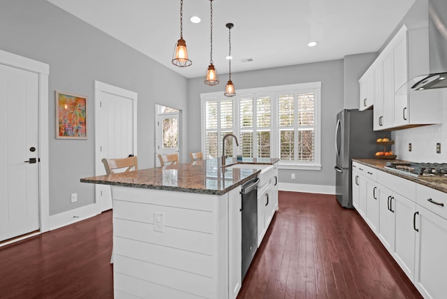 kitchen with a kitchen island with sink, stainless steel appliances, white cabinets, wall chimney range hood, and decorative light fixtures