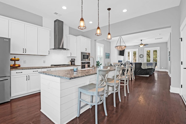 kitchen with stainless steel appliances, wall chimney range hood, a kitchen island with sink, dark stone counters, and ceiling fan