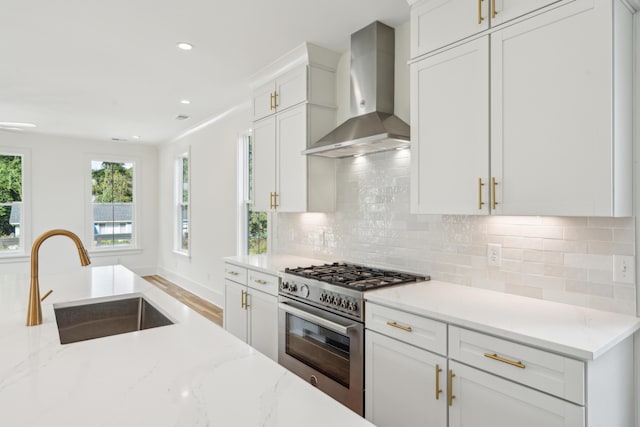 kitchen featuring high end range, white cabinets, wall chimney range hood, sink, and light stone counters