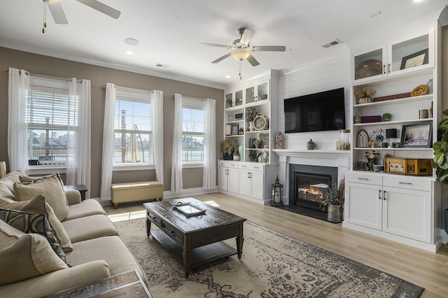 living room with light wood finished floors, a fireplace with flush hearth, crown molding, and ceiling fan