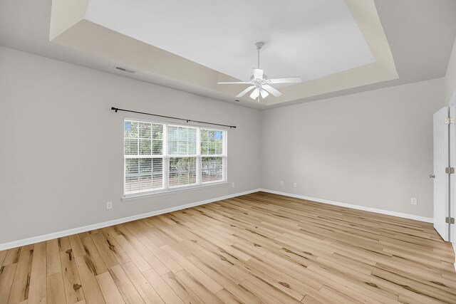 empty room featuring a tray ceiling, ceiling fan, light wood-style floors, and baseboards