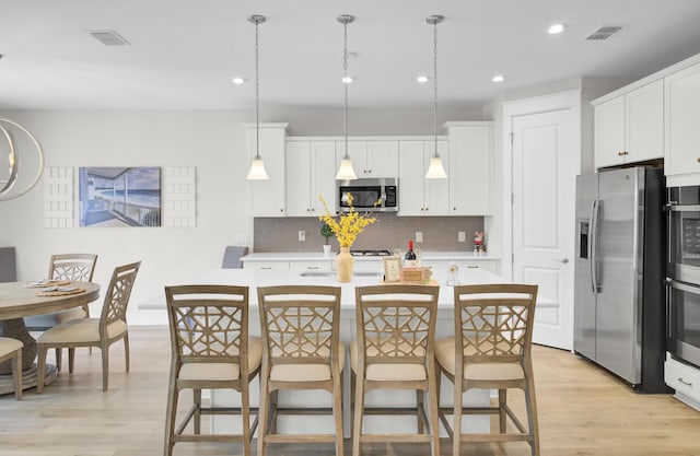 kitchen featuring appliances with stainless steel finishes, an island with sink, white cabinets, decorative light fixtures, and light wood-type flooring