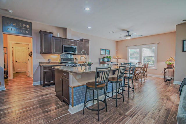 kitchen featuring light stone counters, dark brown cabinets, a kitchen island with sink, ceiling fan, and dark hardwood / wood-style floors