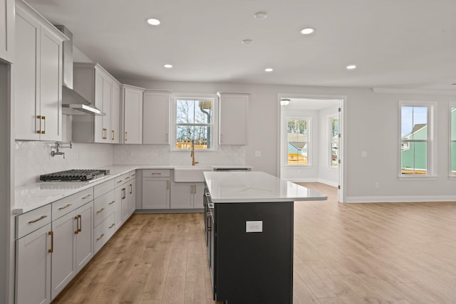 kitchen with light wood-style flooring, light stone counters, wall chimney range hood, stainless steel gas cooktop, and backsplash