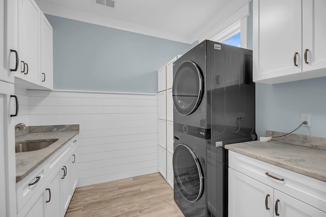 clothes washing area with light wood finished floors, ornamental molding, stacked washer and dryer, cabinet space, and a sink