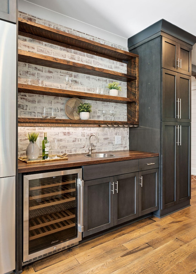 bar with light wood-type flooring, wine cooler, sink, stainless steel refrigerator, and dark brown cabinetry